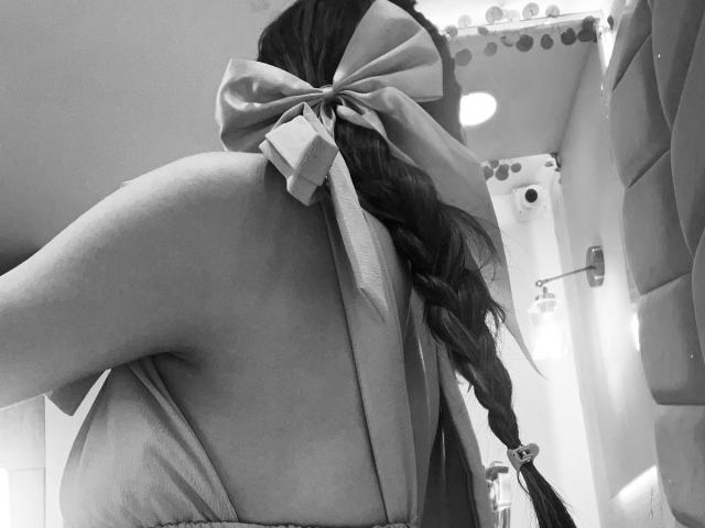 AfroditaWild's back with a large bow on her braid, exposing her bare shoulder and upper back, in a grayscale photo. Her ass is slightly visible from the side.