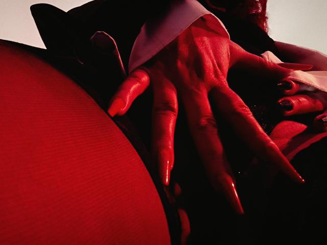 De hand van AmberSunshine, verlicht met rood licht, met lange, glanzende nagels, spreidt haar sappige billen, waardoor een stukje van haar natte, glanzende geslachtsdeel zichtbaar wordt. Haar donkere, gladde huid contrasteert met de rode verlichting..