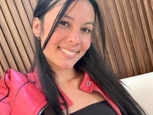 ViolettClay, con cabello negro largo, sonriendo, vestida con una chaqueta roja sobre una blusa negra, mostrando su linda nariz y sus pequeños senos, tiene una mejilla rosada y perforaciones en las orejas..