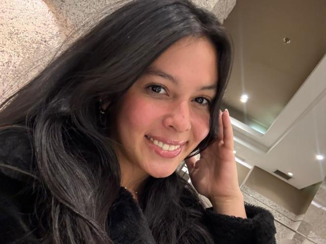 ViolettClay, con lunghi capelli neri, sorridente, indossa un cappotto nero e peloso, ha una mano vicino al viso, occhi marroni e pelle chiara, appare carina e sensuale..