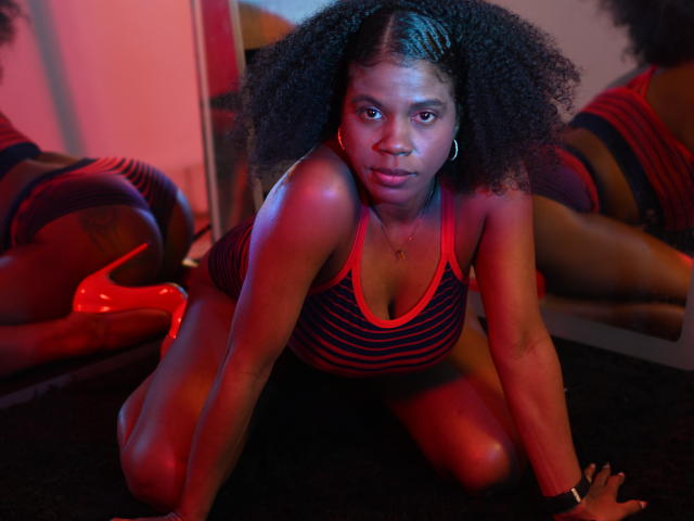 SarahiPusey op handen en voeten, met een tanktop met rode strepen, zwarte shorts en rode hakken, met krullend haar, kijkend naar de camera, reflectie in de spiegel, rode en blauwe verlichting..