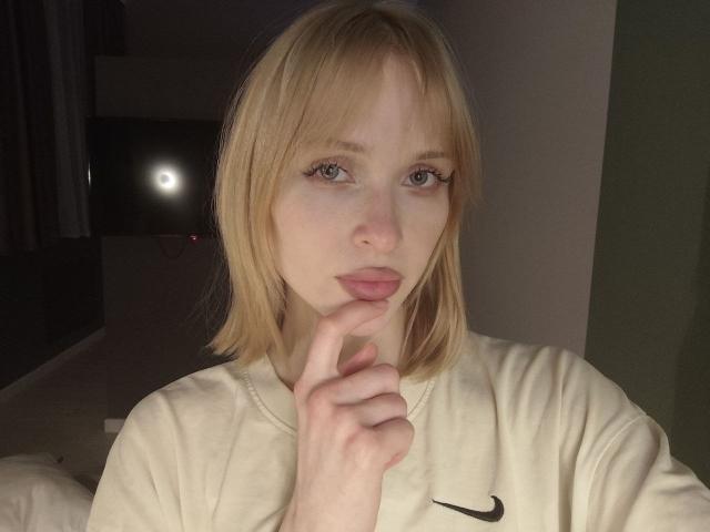 AnnettePantano with blonde bob, blue eyes, and pale skin, wearing a white Nike shirt, has finger on chin, looking at camera, soft light, slightly pouty lips.