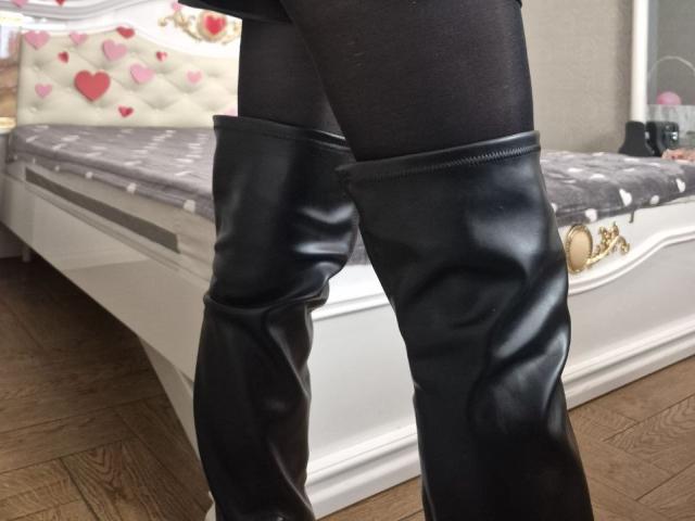 As botas de couro pretas e brilhantes, que chegam até a coxa de Anshante, estão ao lado de uma cama com cabeceira branca e estofada, e um cobertor cinza com padrão de corações..