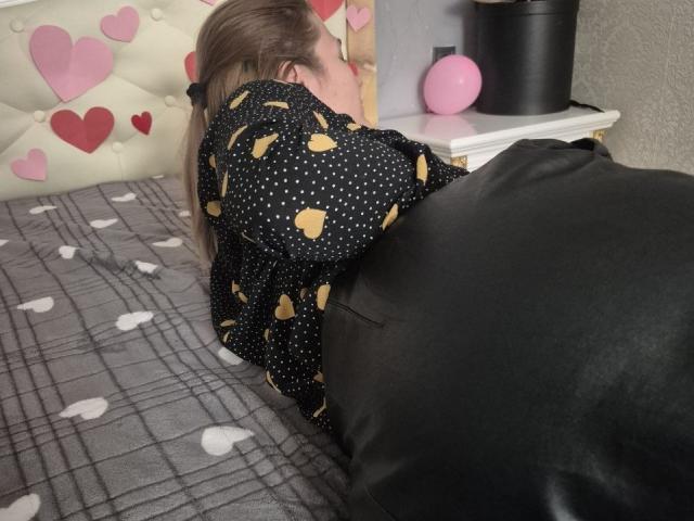 Anshante, con cabello rubio, acostada boca abajo sobre una cama, vestida con una camisa negra de lunares con corazones amarillos, cubriendo sus glúteos con una almohada negra..