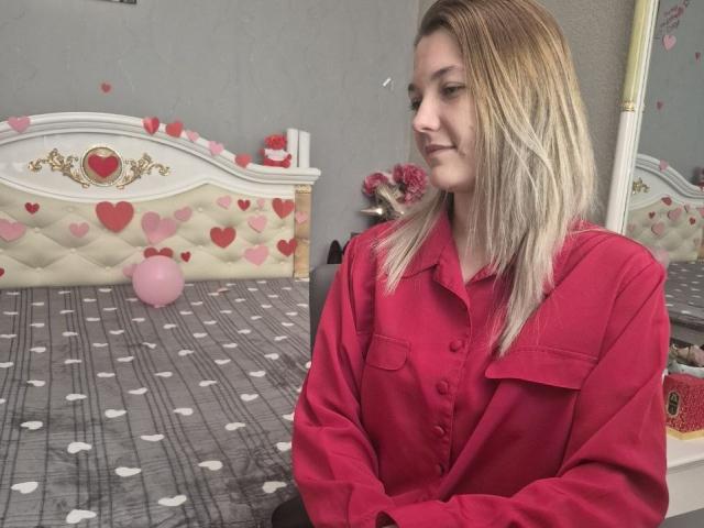 Anshante, con cabello rubio, vestida con una camisa rosa de botones, está sentada en una cama con ropa de cama estampada con corazones y lunares, mirando hacia un lado con una ligera sonrisa..