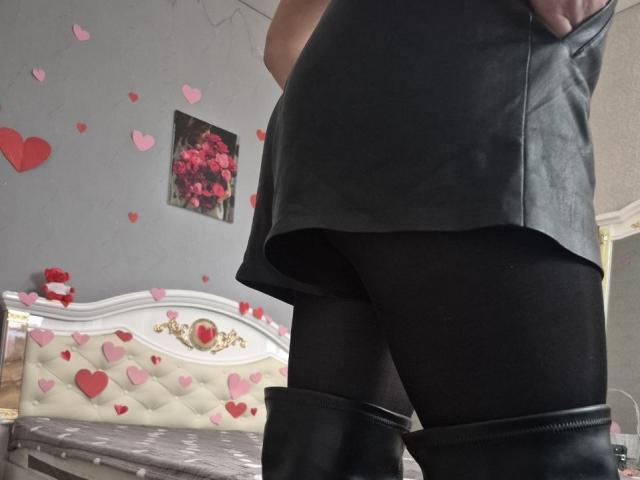 Anshantes schwarzer Lederrock, schwarze Strumpfhosen und schwarze Kniehohe Stiefel. Anshantes Gesäß ist sichtbar und leicht erhöht. Rote Herzverzierungen an der grauen Wand. Ein Gemälde einer roten Rose..