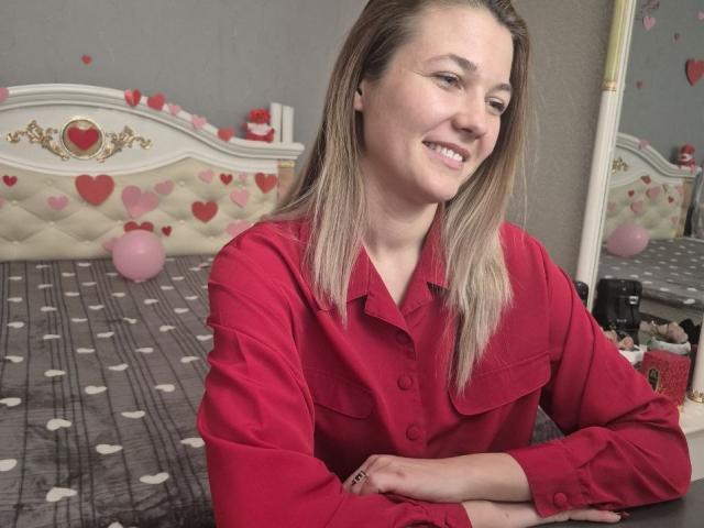 Anshante, con i capelli biondi, indossa una camicia rossa con bottoni, sorride, ha le braccia incrociate e siede sul letto con coperte a motivi di cuori e pois, guarda di lato, illuminazione soffusa..