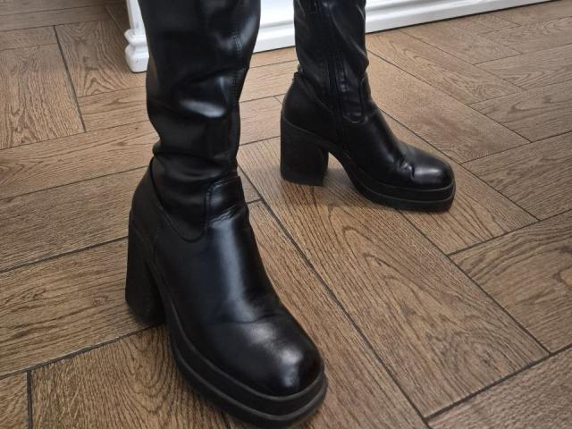 Las botas de cuero negro de Anshante, con tacones negros gruesos, están sobre un suelo de madera. Las botas son brillantes y ligeramente desgastadas, mostrando signos de uso. Los pantalones de cuero negro de Anshante son parcialmente visibles..