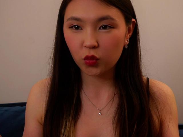 MariLerew with long black hair, red lipstick, and earrings, puckering her lips, wearing a necklace, with visible cleavage, against a plain background.