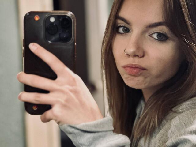 BrigetteLaurie with nose ring, holding phone, taking selfie, pouty lips, brown hair, green eyes, grey shirt, right hand, iPhone, focused expression, soft lighting, realistic photograph.