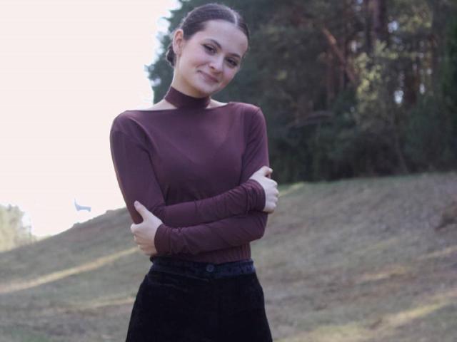 HelgaGriffiths está de pie, con los brazos cruzados y sonriendo, vestida con un jersey de cuello alto color burdeos y pantalones negros, en un área boscosa y soleada. Sus pechos son pequeños y tiene la piel clara..