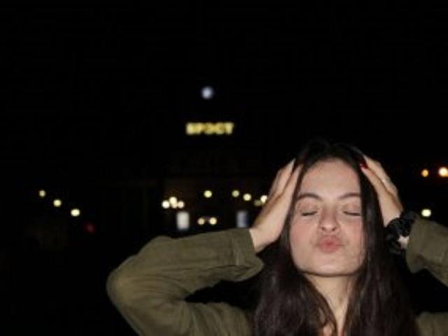 HelgaGriffiths, con las manos en el cabello castaño oscuro, los ojos cerrados y los labios fruncidos, vestida con una camisa verde oliva, en una toma nocturna con luces de la ciudad visibles y una luna..