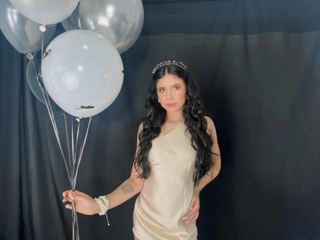 MiluSky, con lunghi capelli neri ondulati, indossa una fascia argentata e un abito beige di seta, tiene in mano palloncini bianchi e argentati, ha dei tatuaggi sul braccio sinistro e un braccialetto bianco..