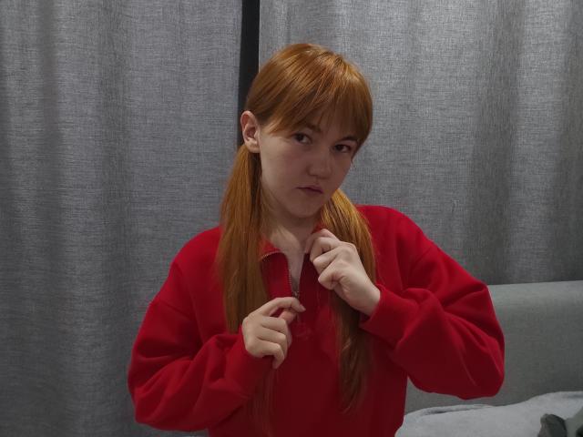 SheritaChieng, con i capelli rossi, indossa un maglione rosso, giocherellando con il bottone della camicia, appare leggermente confusa. Ha un seno piccolo e un incarnato chiaro..