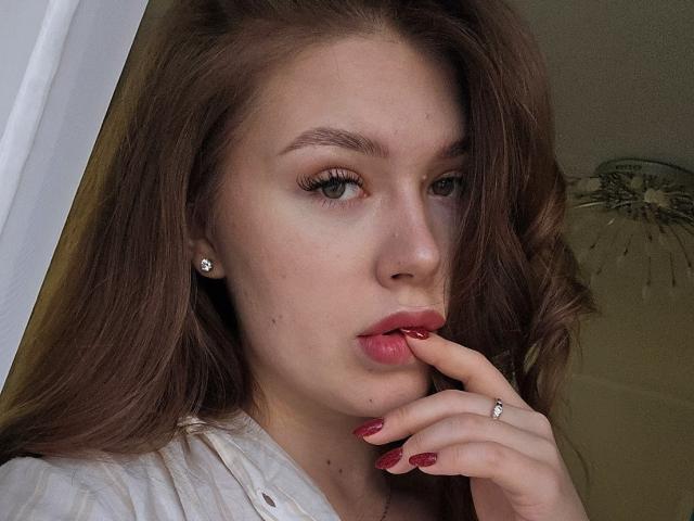 Chrissy Barajas met lang bruin haar, volle rode lippen, een vinger in haar mond, gekleed in een witte blouse, met diamanten oorbellen, een ring, rode nagels en een lichte glimlach..