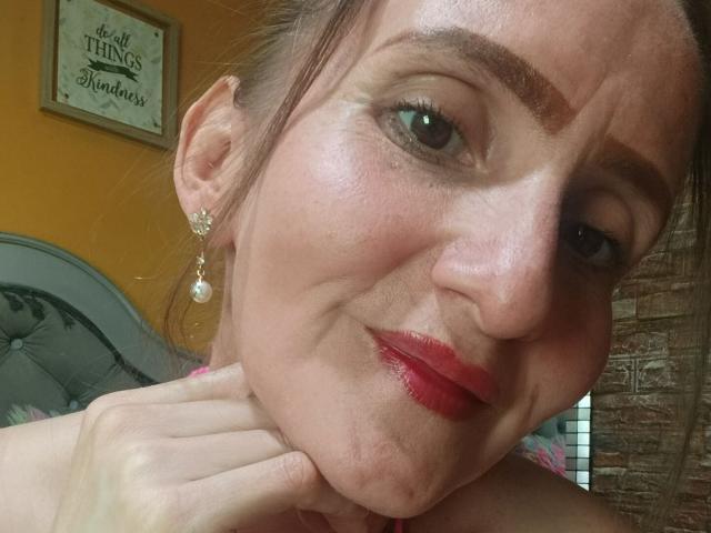 Das Gesicht von RubiAnderson, leicht zusammengekniffen mit einem verschmitzten Lächeln, zeigt roten Lippenstift, Ohrringe und eine Hand, die ihren Hals berührt. Sie hat braue Augenbrauen, grüne Augen und einen hellen Hautton..