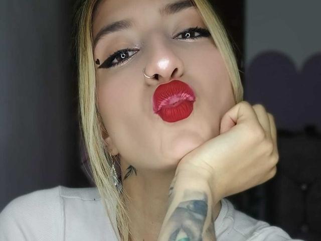 Kaiix mit blonden Haaren, rotem Lippenstift, einem Nasenring und Tätowierungen, zieht ihre Lippen zusammen. Sie berührt ihr Kinn mit einer tätowierten Hand und schaut in die Kamera..