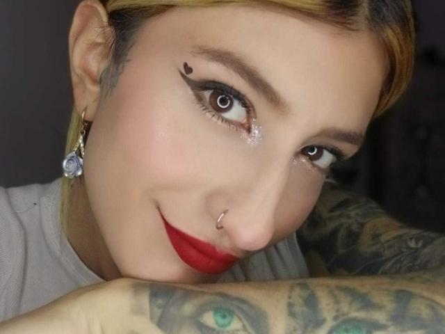 Kaiix hat Tattoos an ihrem Arm, einen Nasenring, schwarzen Eyeliner mit einem Punkt und roten Lippenstift. Ihre Augen sind braun, und sie lächelt mit blonden Haaren..