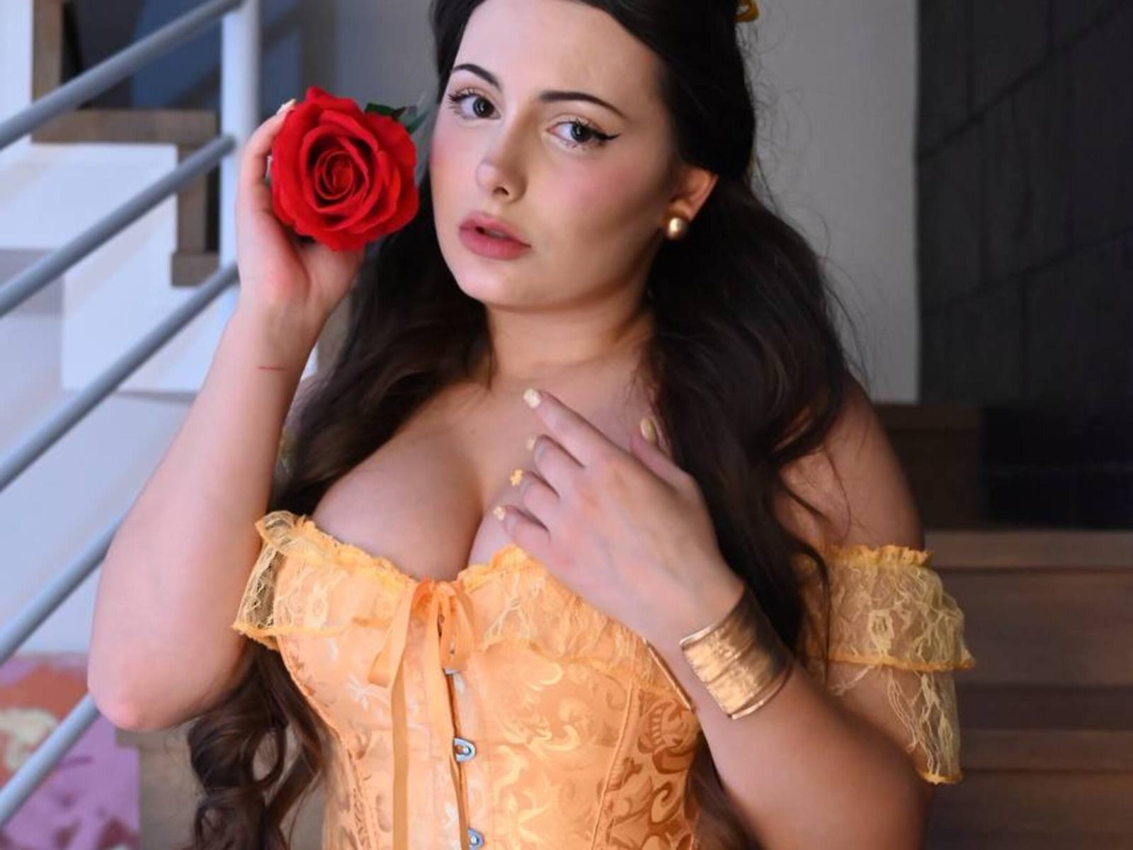 Hildegard Hoppel mit großen Brüsten, die eine rote Rose in der Nähe ihres Gesichts hält, trägt ein orangefarbenes Spitzenoberteil mit einem Schulterteil, hat langes, dunkles Haar, ein goldenes Armband und Ohrringe und sieht niedlich aus..