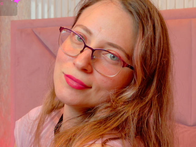 AmbeerWood with glasses, pink lipstick, and brown hair, smiling slightly. She has a nose piercing and a pink shirt. Her blue eyes are visible. Her tits are not shown.