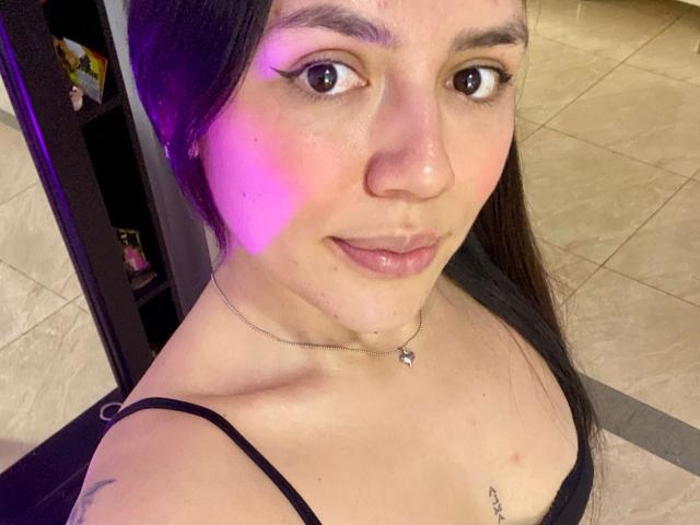 SophiaMarx has pink light on her cheek, black hair, brown eyes, and a black strap top showing her small tits and a nipple piercing. She's smiling with a silver necklace.