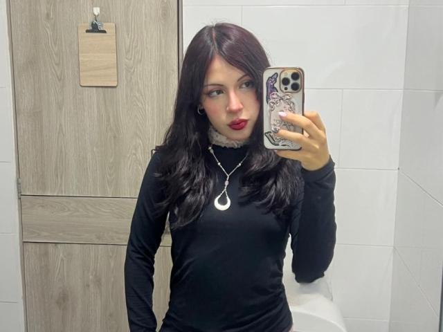 Valery Beaumont ha un rossetto rosso scuro, una camicia nera a maniche lunghe, una collana d'argento con un ciondolo a forma di mezzaluna e tiene in mano un telefono con una custodia a motivi viola e bianchi, in un bagno..