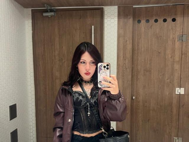 Valery Beaumont, com cabelo ruivo escuro, vestindo uma jaqueta roxa, um corpete preto com cadarços e um colar, tirando uma selfie no espelho, mostrando seus seios e uma pequena tatuagem no pescoço..