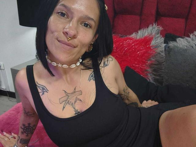 AmelieBlake, with black hair, nose ring, and tattoos, sits on a bed wearing a black tank top and shell choker, showing small tits and a slight smile.