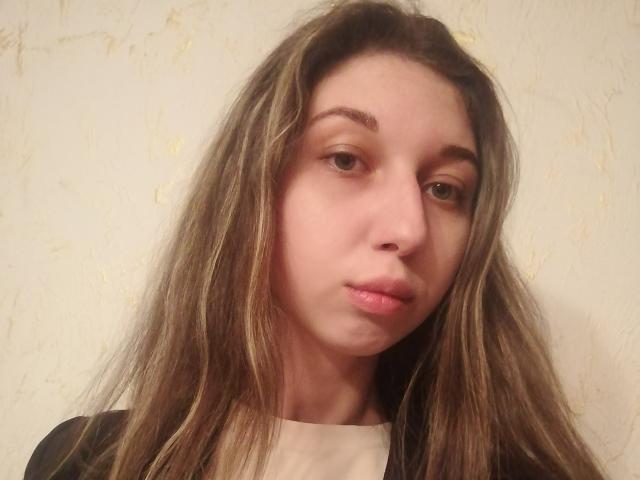 Sanella tiene el cabello largo y rubio oscuro, ojos marrones claros y un rostro ligeramente fruncido con labios rosados. Lleva una blusa negra y blanca..