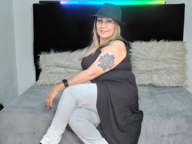 MatureYayita met blond haar, een zwarte mouwloze jurk, witte broek, een zwarte pet, een roos tatoeage op haar arm, lachend, zittend op een grijs bed met pluizige witte kussens..