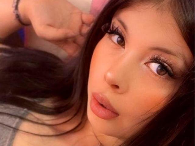 Het gezicht van JessyKim, met bruine ogen, lange wimpers en roze lippen, kijkt opzij met een lichte frons, bruin haar, en een grijze top, wat een schattige, sexy uitstraling geeft..