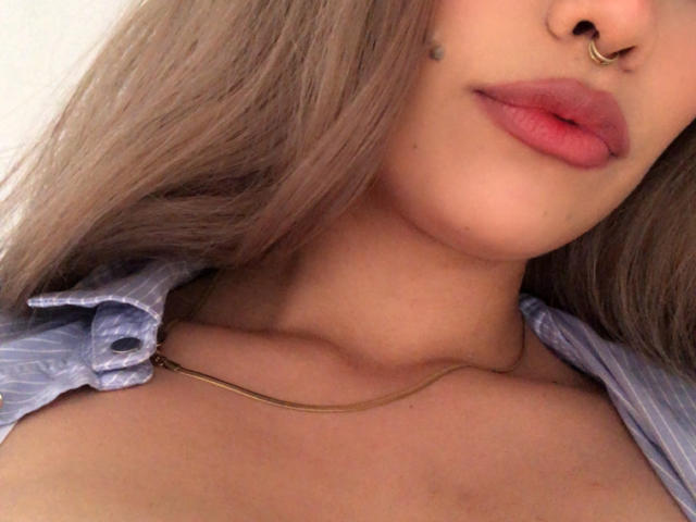 Ilayna's face with pink lipstick, gold nose ring, and small beauty mark. She's wearing a blue striped shirt, and a thin gold chain necklace. Her tits are slightly visible.