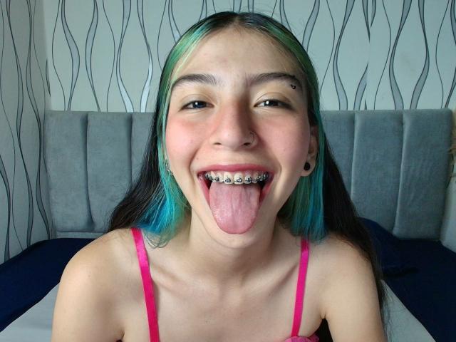 AbbyHillsI has green and blue dyed hair, black nose ring, and is sticking out her tongue with metal grills in her teeth. She's wearing a pink top.