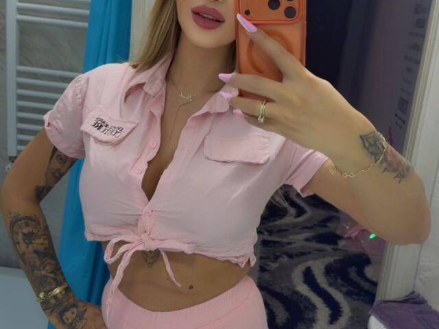 NicolSNaya trägt ein pinkes, hochgekrempeltes Hemd, das ihre Brüste zeigt, pinke Hosen, hat Tattoos, pinke Nägel und hält ein orangefarbenes Handy, mit dem sie ein Selfie vor einem Spiegel macht. Ihre Lippen sind pink gestrichen..