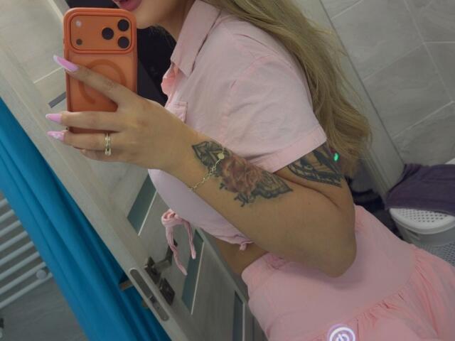NicolSNaya con labbra rosa, che tiene in mano un telefono arancione, con unghie rosa, un outfit rosa, mostra un braccio tatuato, capelli biondi, un anello al dito, in un selfie allo specchio del bagno..