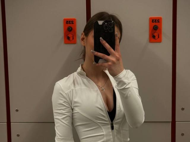 LesterKassner with tits visible in a tight white zip-up shirt, holding a phone in front of her face, standing in front of locker doors with orange buttons.