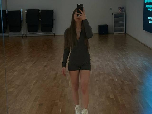 Lester Kassner, in piedi, sta scattando un selfie con uno specchio, indossa una maglia a maniche lunghe grigio scuro e pantaloncini, scarpe da ginnastica bianche, ha capelli castani lunghi, tiene in mano un telefono, pavimento in legno, parete neutra..