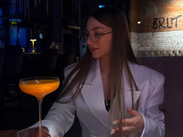 Lester Kassner, con lunghi capelli castani, occhiali, camice da laboratorio bianco, che tiene in mano un cocktail arancione, seduto a un tavolo scuro del bar, che guarda di lato, con la mano destra che tiene il bicchiere del cocktail..