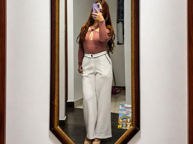 VicluX avec de longs cheveux bruns, un haut rose à manches longues, un pantalon blanc taille haute, tenant un téléphone, autoportrait devant un miroir, debout, cadre marron, murs blancs, sol beige, talons marron..