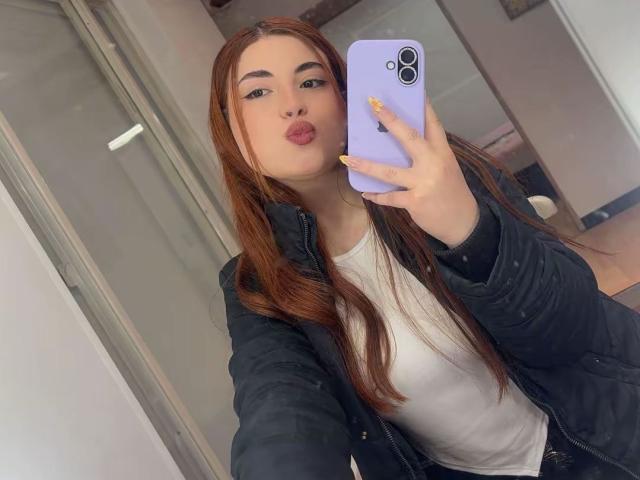 VicluX, con lunghi capelli rossi, labbra tumefatte, indossa una giacca nera e una maglietta bianca, sta scattando un selfie con un telefono viola, tenendolo sollevato verso il viso..