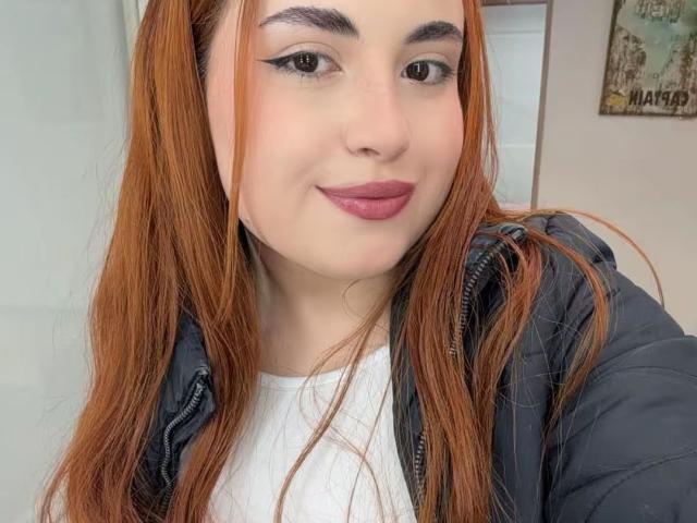 VicluX has long red hair, black eyebrows, and wears dark lipstick with black eyeliner. She's in a gray jacket over a white shirt, smiling softly.