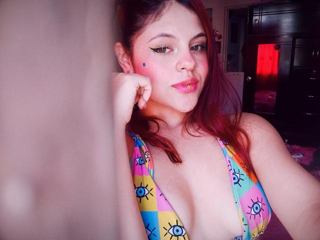 Sophie Hill mit roten Haaren, einem bunten Bikini mit Augenmuster, einem blauen Punkt auf der Wange, einem Nasenring, pinkem Lippenstift und Eyeliner, lehnt an einer Wand, lächelt und zeigt ihre Brüste..