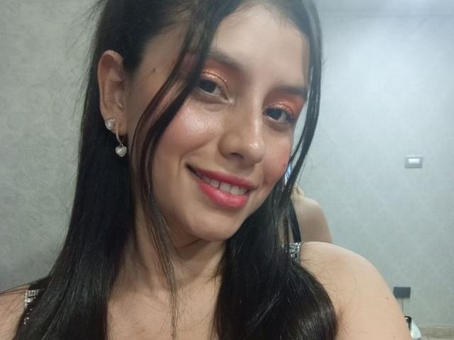 MadissonFontaine tem sombra laranja brilhante, batom rosa e um sorriso, com cabelos pretos longos, brincos de diamante e uma blusa brilhante, mostrando seu rosto adorável e pele lisa..