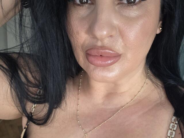 MisttresRbk's face with shiny, glossy pink lips, dark black hair, and a gold necklace. Her skin is slightly tan, and she has a serious, sultry expression. Boobs visible.
