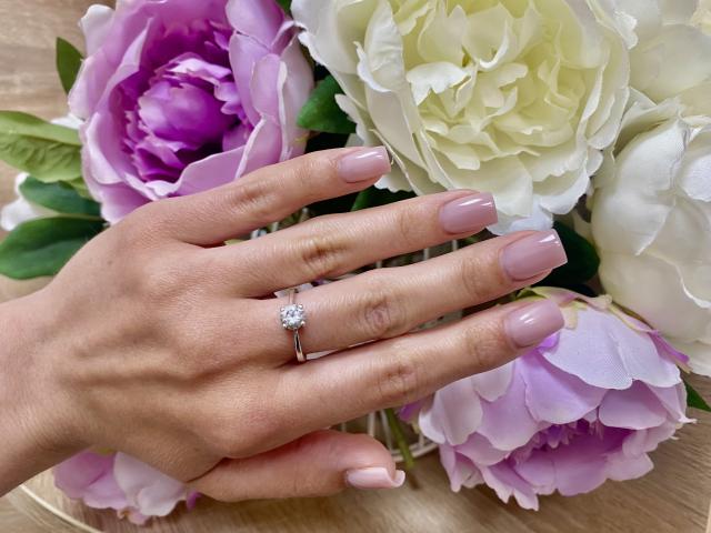 La main d'OliviaGranda, avec son vernis à ongles rose et son anneau en diamant, repose sur un bouquet de pivoines blanches et violettes. Ses doigts sont légèrement recourbés, mettant en valeur sa manucure..