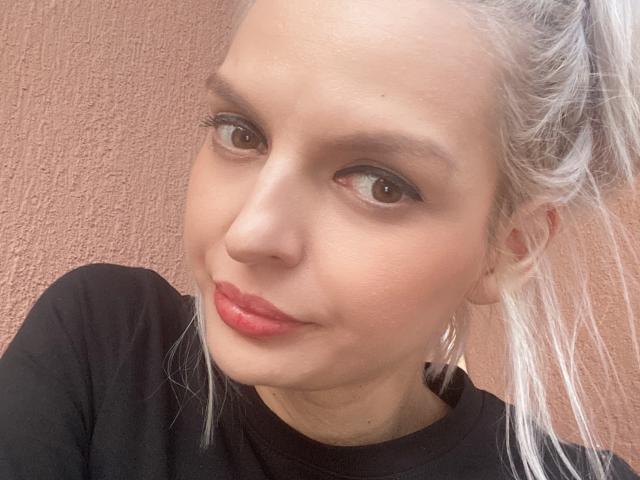 DouceAmelia has pale skin, brown eyes, red lipstick, and white hair in a ponytail, wearing a black shirt, with a subtle smile and small hoop earrings.