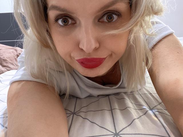 DouceAmelia with blonde hair, red lipstick, and a white shirt, lying on a bed with a geometric-patterned sheet, looking at the camera with a slight smile.