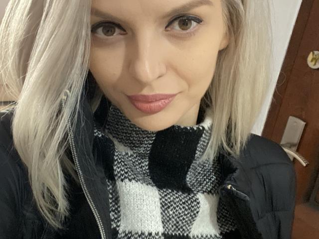 DouceAmelia with blonde hair, blue eyes, and pink lips, wearing a black and white checkered scarf, and a black jacket, looking directly at the camera, smiling slightly.