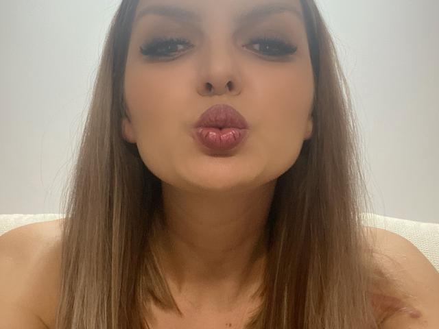 DouceAmelia's face with pouty pink lips, long brown hair, and heavy black eyeliner, making a kissy face in a selfie, showing off her slightly parted, glossy lips.