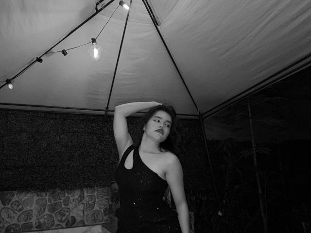 Black-and-white photo of AlexaMissFortune with long dark hair, wearing a sparkly, one-shoulder dress, hand on head, looking up at hanging light bulbs. Boobs visible under dress.